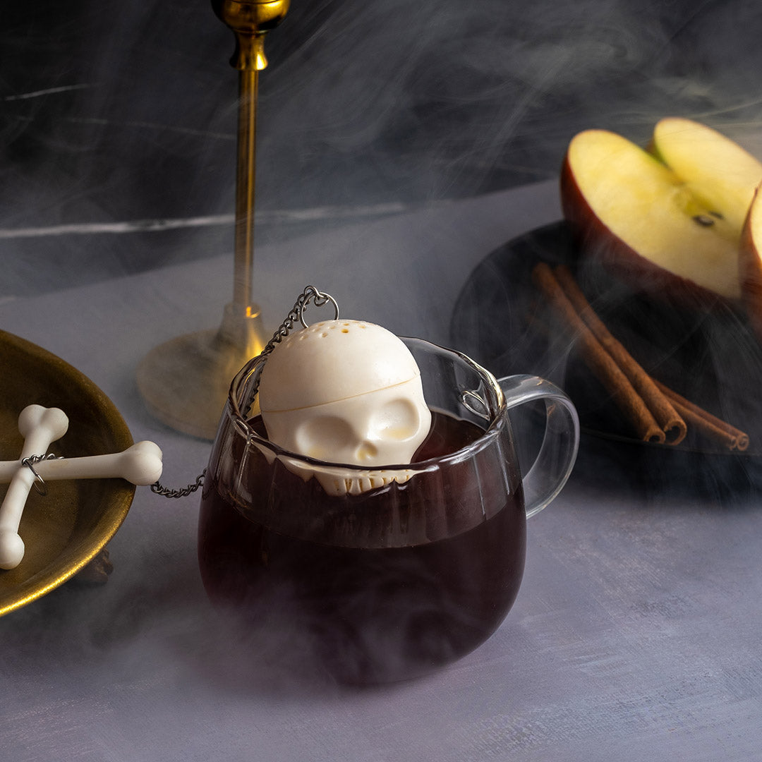 A skull-shaped teapot resting in a glass mug filled with dark coffee, surrounded by moody props including a brass candlestick, sliced apple, cinnamon sticks, and a bone-shaped object on a smoky gray surface.